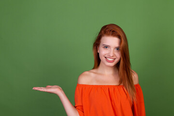 Young red hair woman in casual orange blouse on green background smile  and laugh holds empty space on hand