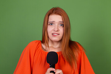 Young red hair woman in casual orange blouse on green background holding microphone worried shy to...