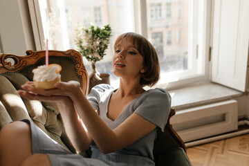 Lifestyle portrait of young lady at home. Charming dark-haired girl in blue clothes, holding cake with candle, resting on armchair against white window