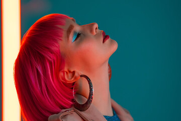 Eccentric woman with pink hair posing near led neon lamp on teal background. Charming odd girl,...