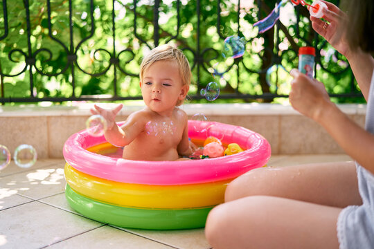 Mom blows soap bubbles, and a little girl catches them while sitting in an inflatable pool