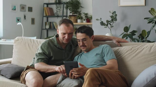 Portrait of two handsome Caucasian men in love spending time together at home sitting on sofa surfing Internet on digital tablet