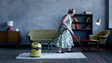 Happy elderly woman listening music on headphones and vacuum cleaning carpet