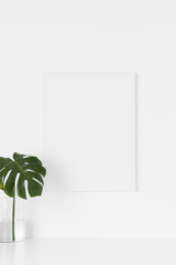 White canvas mockup on the wall with a  monstera leaf in a glass vase.