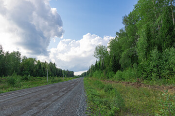 Fototapeta na wymiar The road against the background of blue sky with white clouds on a bright sunny summer day. Part of birch trees with green foliage. Overall plan. Scenery