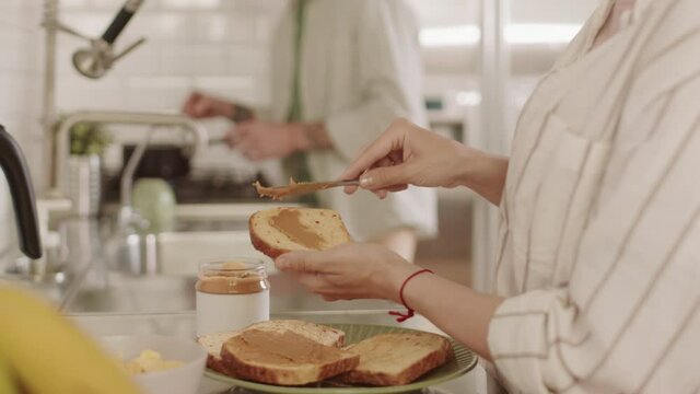 Side view slowmo of cropped unrecognizable person standing by kitchen counter, making toast with peanut butter, blurred man cooking on background