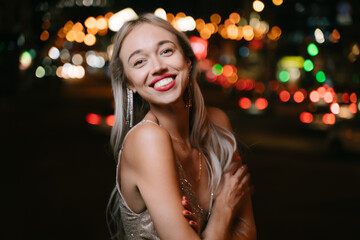 Fototapeta premium satisfied blonde woman smiling looking at camera at summer evening street. Stylish fancy golden cocktail Glided dress and long earrings. Summer party mood. Bokeh lights background. Red lips
