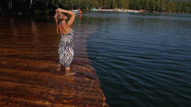 Woman walk on water on pier in sunglasses and a boho silk shawl. Girl rest on a flood wood underwater dock. The pavement is covered with water in the lake. In the background are mountain and a forest.