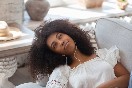 Young black woman relaxing on comfortable couch, enjoying listening to lounge music live via smartphone app, calm without stress, eyes closed, meditation at home concept.
