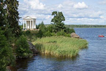 Obraz premium VYBORG, Leningrad REGION, RUSSIA-JULY, 19, 2021: view of the white temple of Neptune and the green foliage of trees on a summer cloudy day in the Monrepos Park and the beautiful shore and blue water