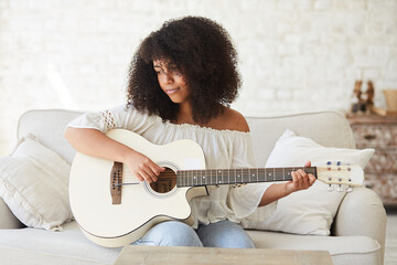 Young afro american woman relaxing with music and playing acoustic guitar at home. Mixed race teenager learning to play guitar using online tutorials. Online music concert from home.