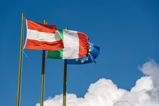 Austrian, Italian and European Union flags with flagpole, blowing togetherness in the wind on a clear blue sky with clouds and copy space.