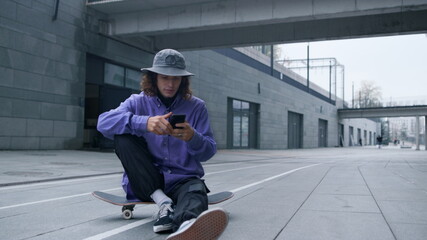 Skater using smartphone outdoor. Hipster sitting on skateboard with phone.