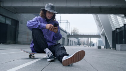 Sporty hipster reading cellphone on skate. Man sitting on skateboard with phone.