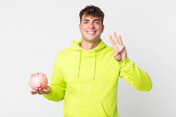 young handsome man smiling and looking friendly, showing number three and holding a piggy bank