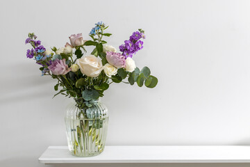Bouquet of hackelia velutina, purple and white roses, small tea roses, matthiola incana, blue iris in glass vase is on the white coffee table .