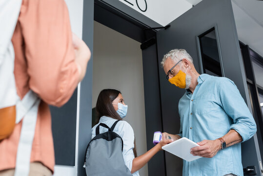 low angle view of teacher in orange medical mask checking temperature of schoolgirl with pyrometer