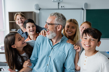 happy multiethnic pupils embracing laughing teacher in classroom