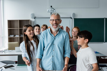cheerful multiethnic classmates near happy teacher laughing with closed eyes in classroom