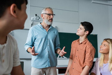 Obraz premium grey haired teacher gesturing while talking to teenagers in classroom