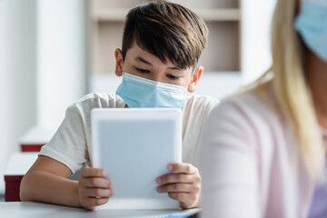 Asian schoolboy in medical mask using digital tablet in classroom