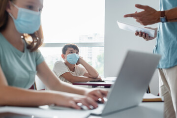 Asian schoolkids in medical mask looking at blurred teacher with digital tablet