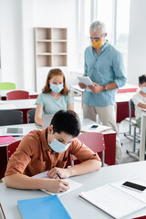 Obraz premium Schoolkid in medical mask writing on notebook in classroom