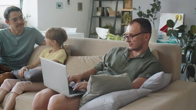 Serious mature man sitting on sofa working remotely on laptop, his husband and daughter sitting next to him chatting about something