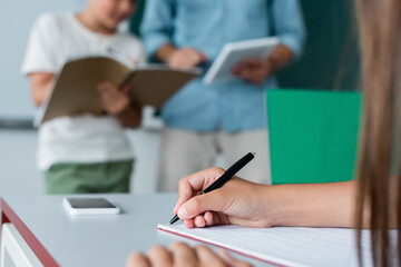 Cropped view of schoolkid writing on notebook near smartphone in classroom