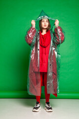 Calm female wearing transparent plastic raincoat and red clothes standing on green background in studio