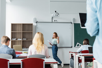 Smiling pupil with notebook talking near erase board and classmates