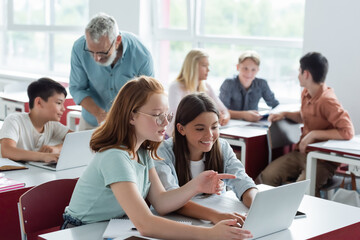 Schoolkid pointing at laptop near teacher and multiethnic classmates