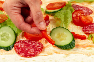 female hand puts fresh chopped vegetables on unwrapped shawarma with sauce, cucumber, lettuce and salami sausage. selective focus. close up
