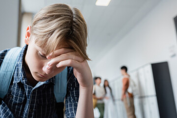frustrated teenager with bowed head near classmates on blurred background