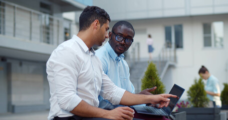 Obraz premium Multiethnic office colleagues using laptop sitting on bench outdoor.