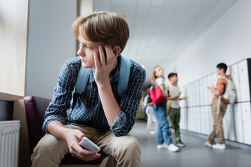 depressed boy with smartphone sitting alone in school corridor near teenagers on blurred background - Powered by Adobe