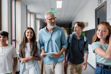 cheerful teacher with digital tablet walking along school corridor with teenagers
