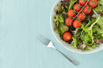 Lettuce leaves and cherry tomatoes in a bowl and a fork on light blue surface, top view with copy space