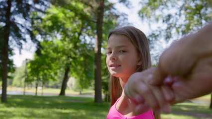 Fototapeta premium Pov shot of beautiful teen girl holding friend hand and looking at camera walking in summer park