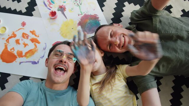 From above view shot of cheerful gay men and their cute child relaxing on floor waving hands after fingerpainting