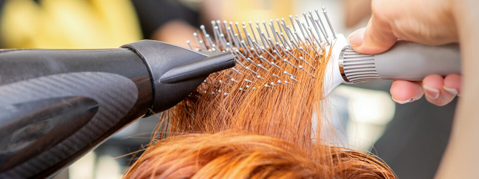Closeup of master's hand with blow-drying and hairbrush blowing female red hair in a salon