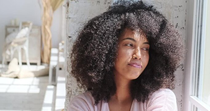 Relaxing smiling young black woman is sitting woman sitting near window, resting, meditating, napping, enjoying a nice stress-free weekend time alone in her home. Pause in remote work.