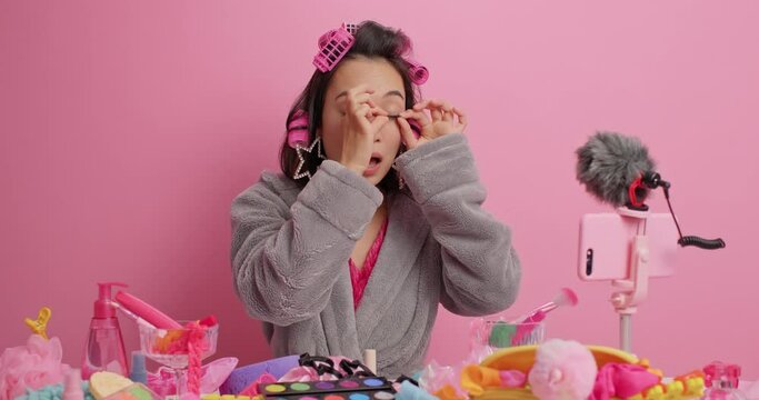 Female beauty blogger records commercial content does visage applies false eyelashes hair rollers and bathrobe shoots masterclass poses at table with cosmetic products isolated on pink wall.