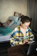 Fototapeta na wymiar Front view of caucasian woman working on laptop from home, in cozy atmosphere, with flowers on the desk. Female wearing round eyeglasses is using smartphone. Her little daughter lie in bed behind