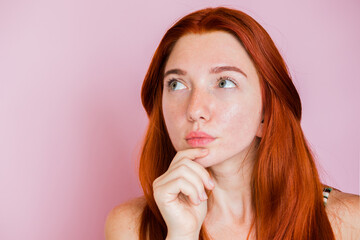 Fototapeta premium A beautiful red-haired girl with natural beauty looks up and is thinking about something. Red-haired girl on a pink background.