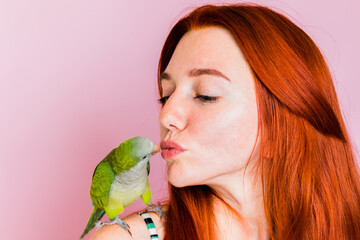 A less-haired girl kisses a green monk parrot who sits on her shoulder on a pink background.