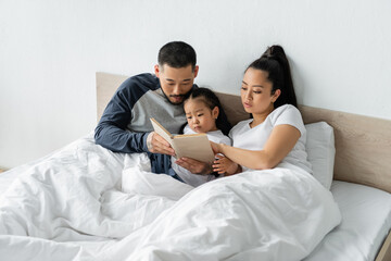 Fototapeta premium asian parents reading book to toddler daughter in bed