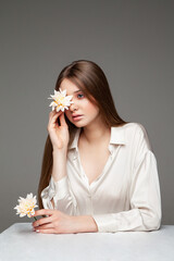 Young female with long brown hair covering eye with natural flower and looking away while sitting at table against gray background