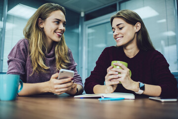 Cheerful female employees with education notepad and smartphone technology for creating content blog plan talking and smiling at table desktop, funny Caucaisan colleagues discussing information