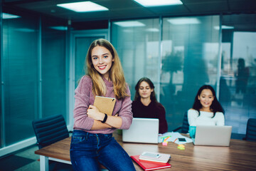 Half length portrait of happy female employee holding education copybook in hands and smiling at camera while sitting at table desktop with blurred colleagues on background, woman with textbook posing